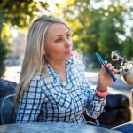 Girl with long blond hair in tshirt sitting at the table outside and smoking electronic cigarette.